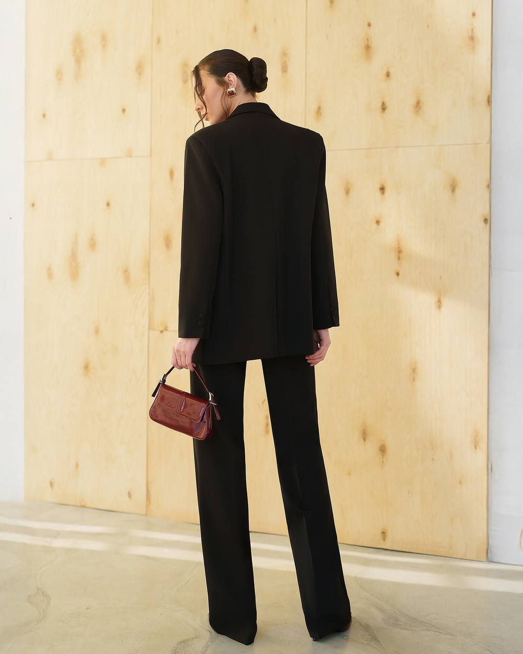 Person wearing a black suit holding a brown handbag against a wooden paneled wall.