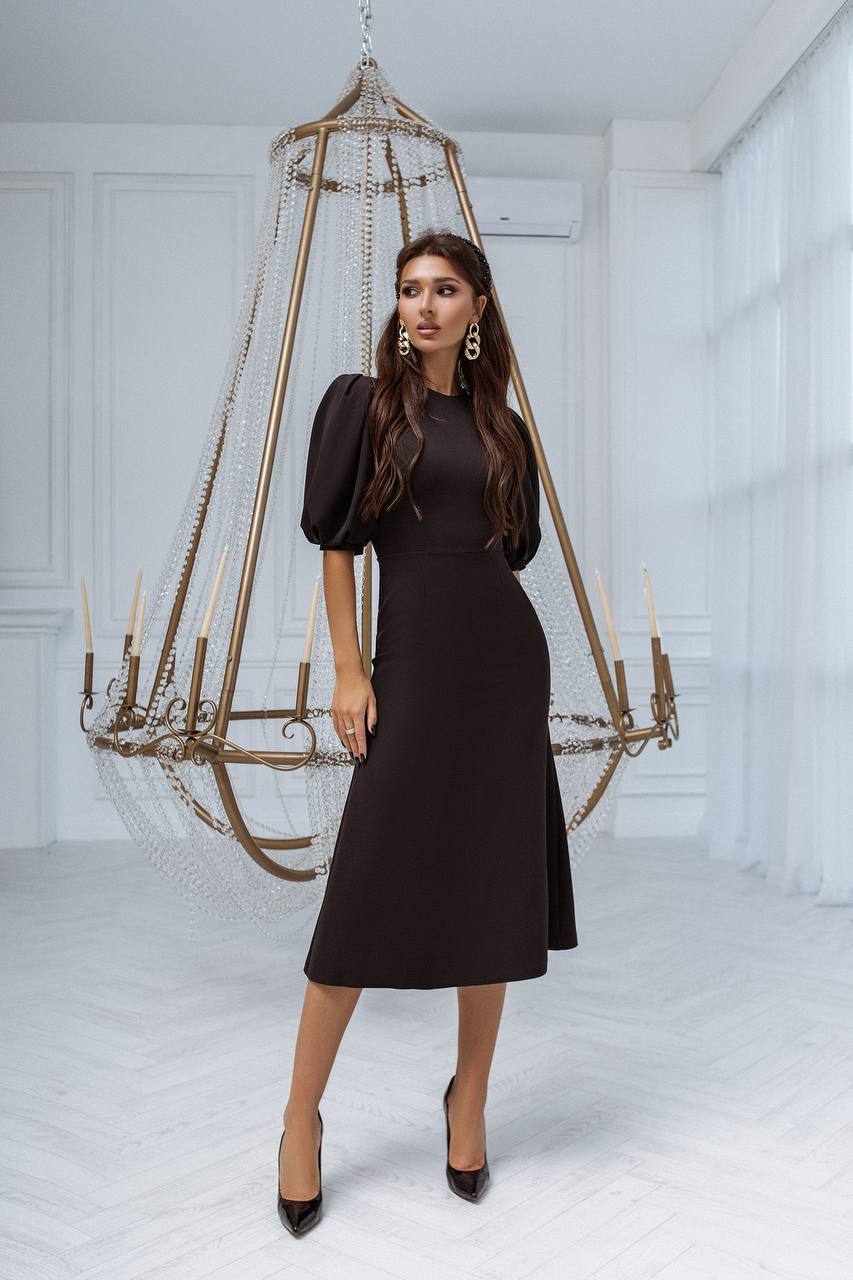 Woman in a black dress standing next to a decorative hanging cage in a white room.