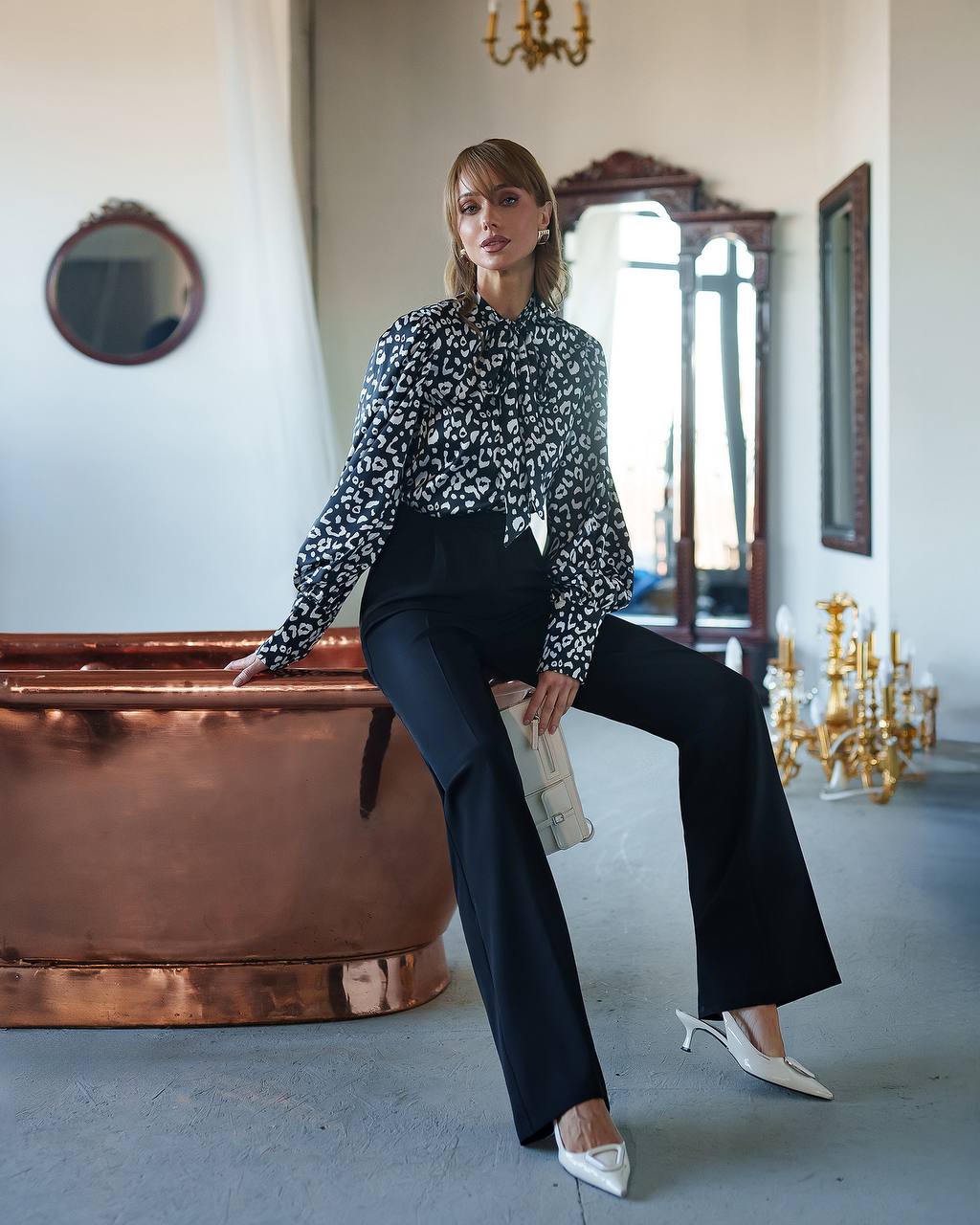 Woman in a floral blouse and black pants sitting in an elegant room with a chandelier.