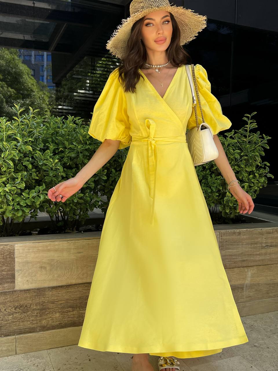 Woman in a yellow dress and straw hat standing outdoors with greenery in the background