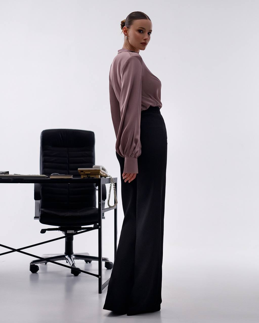 Woman in a pink blouse and black pants standing in an office setting with a desk and chair.