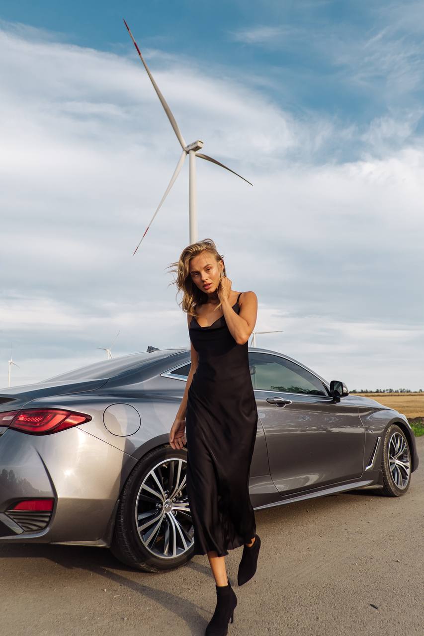 Woman in a black dress standing next to a silver car with a wind turbine in the background