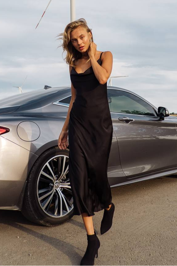 Woman in a black dress standing next to a car with a cloudy sky background
