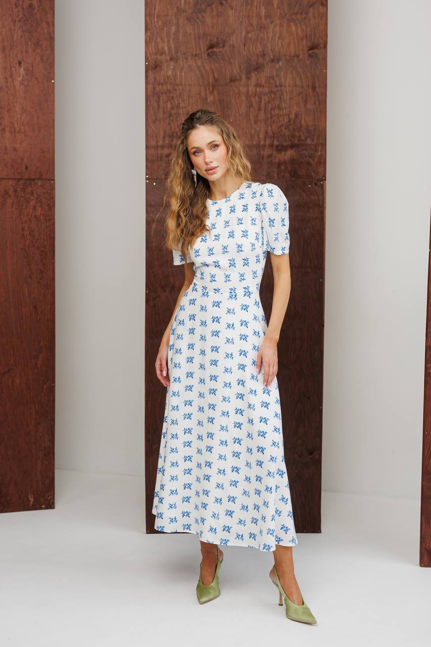 Woman wearing a white dress with blue patterns standing against a wooden door.