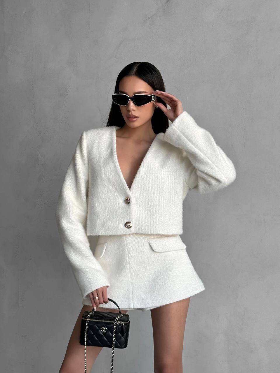 Woman in a white outfit with sunglasses and a black handbag against a gray background
