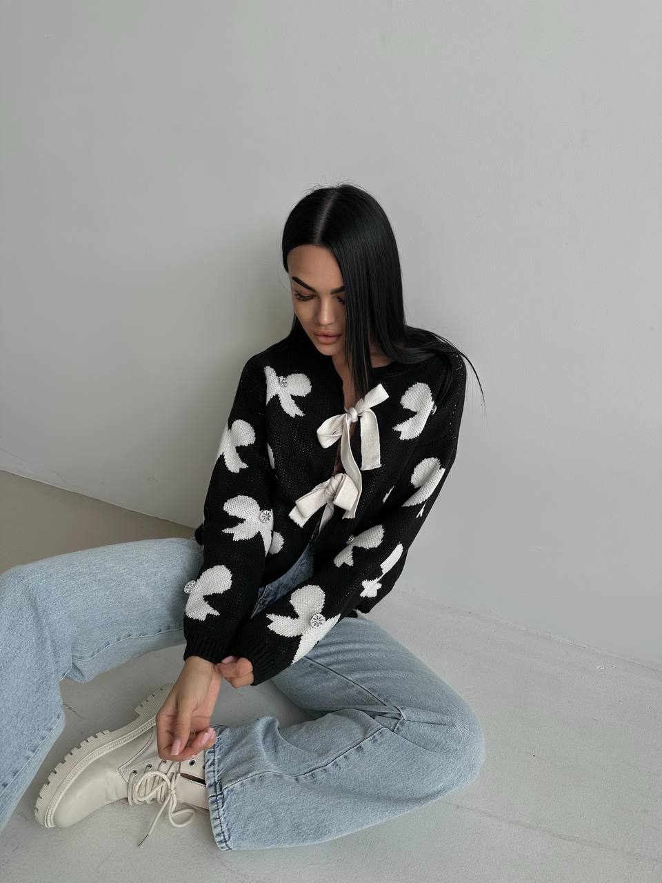 Woman wearing a black sweater with white patterns, light blue jeans, and white sneakers on a plain background.