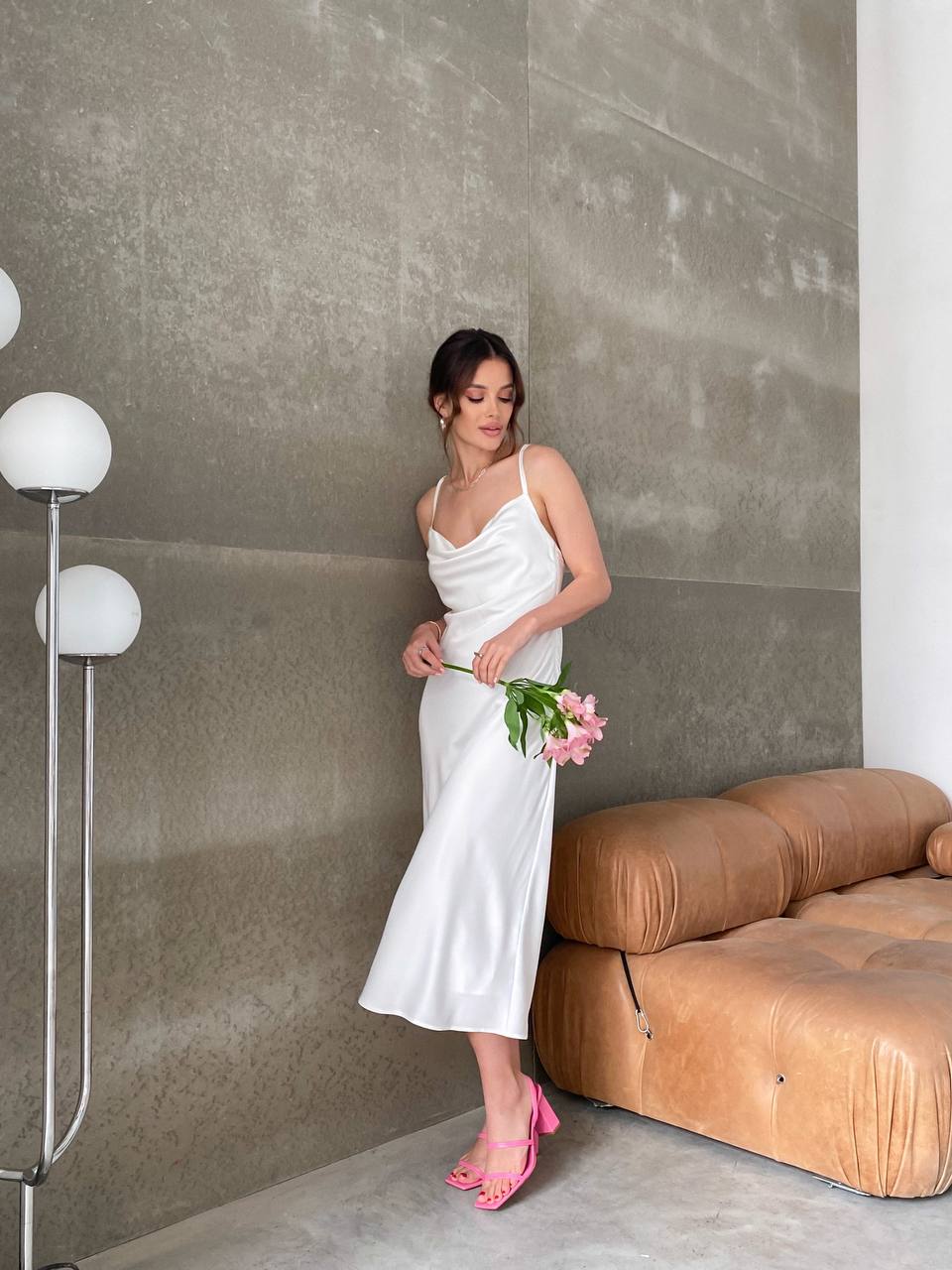Woman in a white dress holding pink flowers in a modern room with a brown sofa and gray wall.