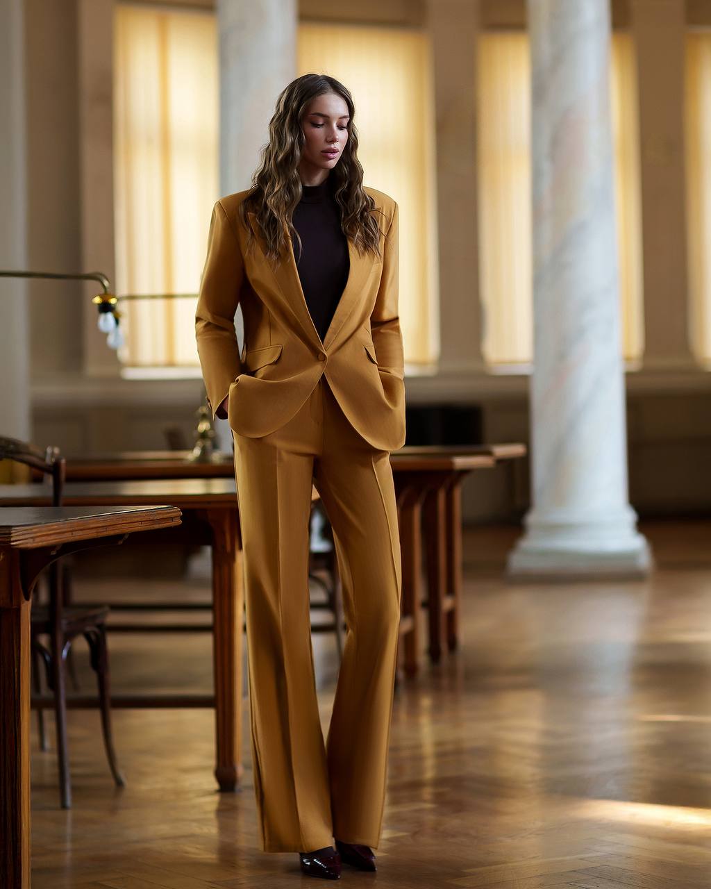 Woman in a mustard suit standing in an elegant room with columns and wooden tables.