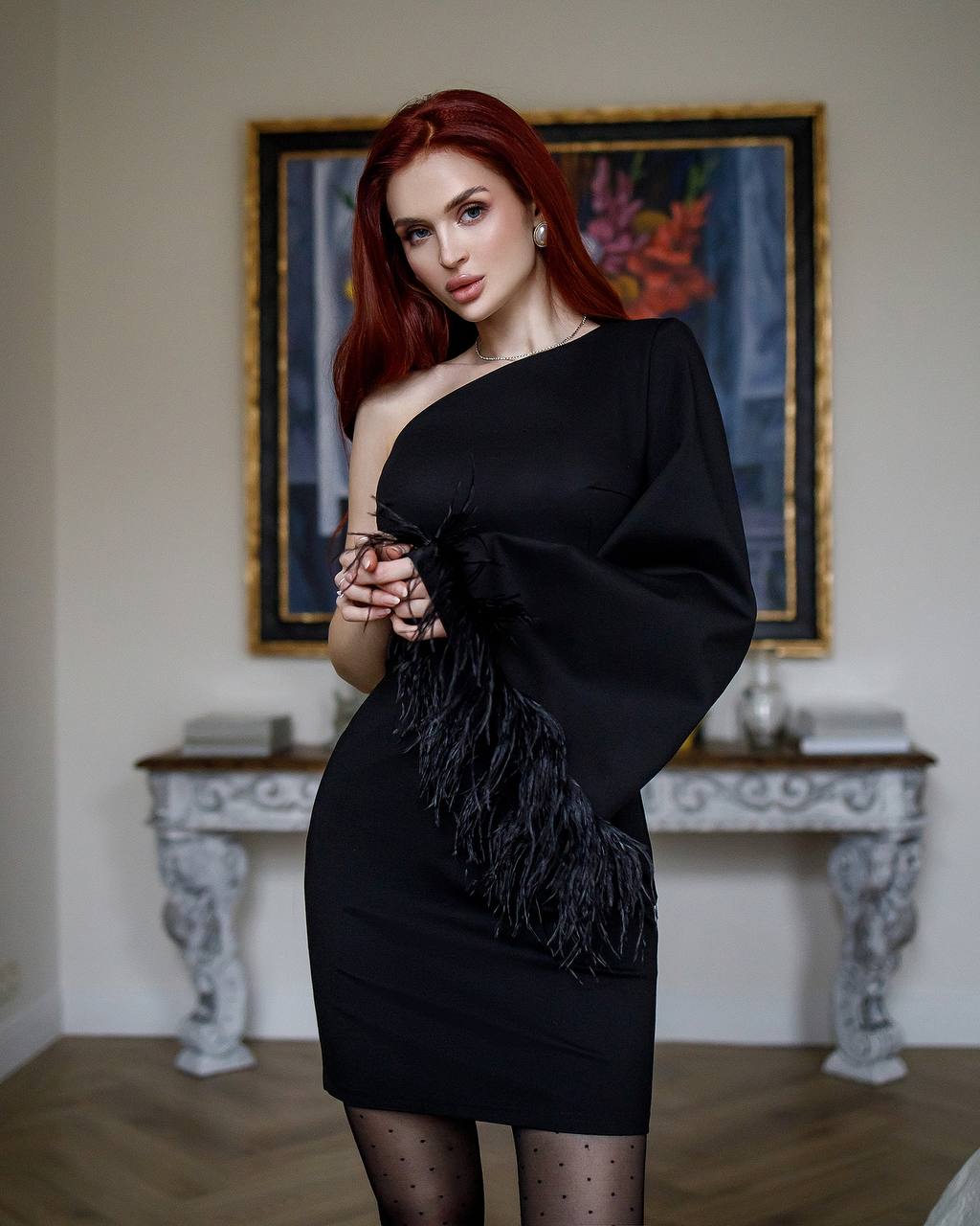 Woman in a black one-shoulder dress with feather details standing in an elegant room.