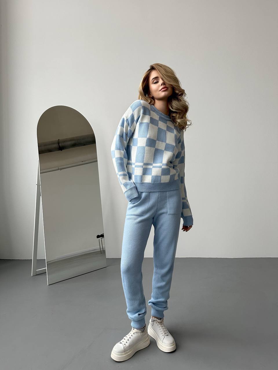 Woman wearing a blue checkered sweater and pants standing in front of a mirror.