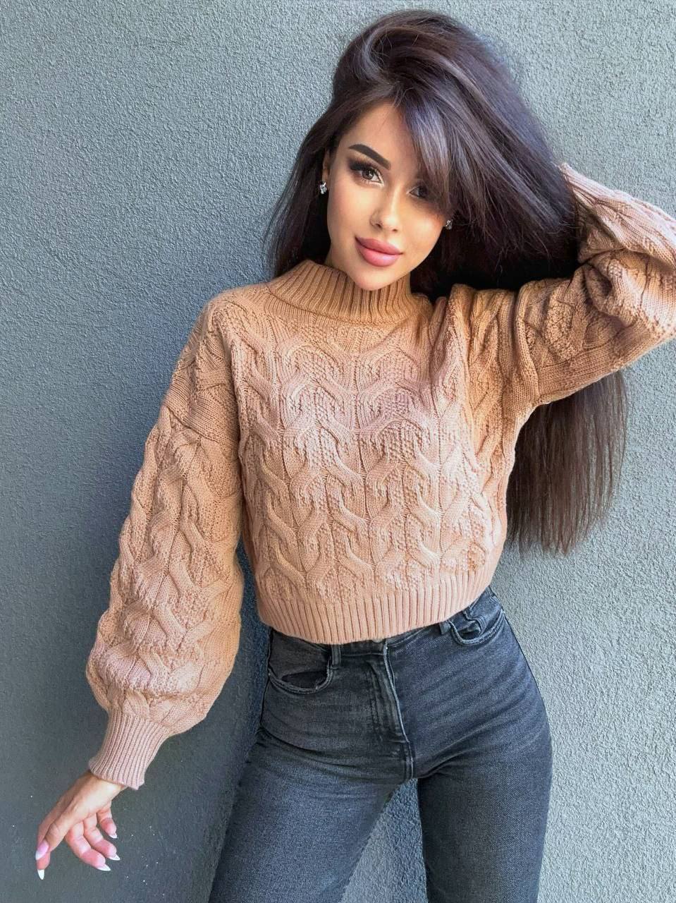 Woman wearing a beige cable knit sweater and dark jeans against a gray wall.