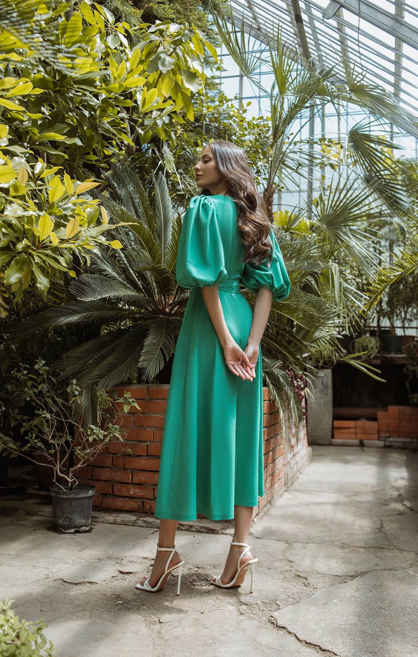 Woman in a green dress standing in a greenhouse filled with plants.