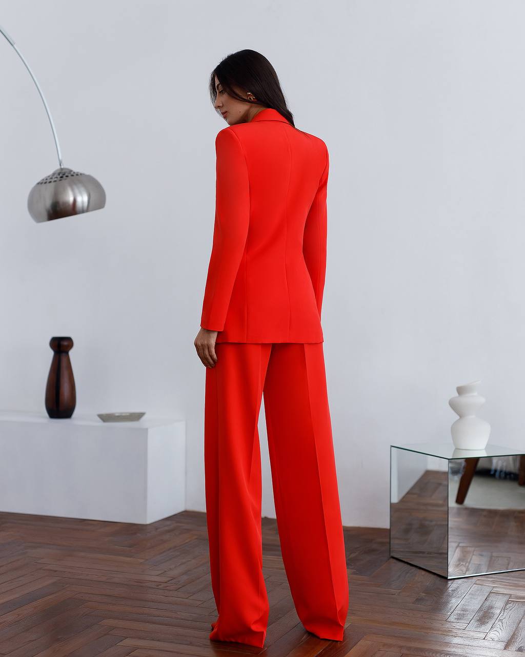 Person wearing a bright red suit standing in a minimalistic room with a white wall and wooden floor.