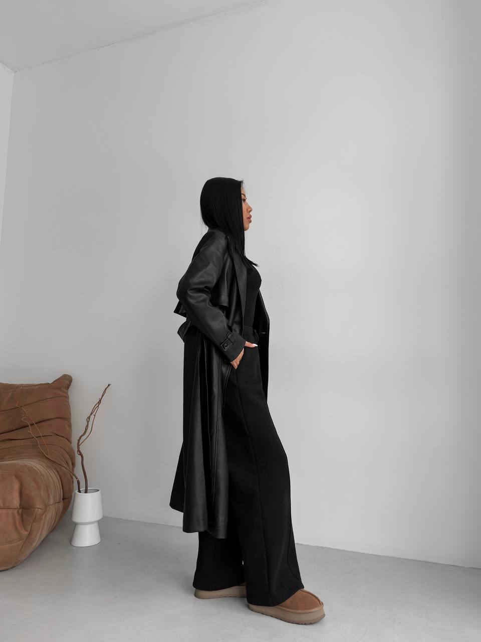 Person wearing a black outfit standing in a minimalistic room with a white wall and brown cushion.