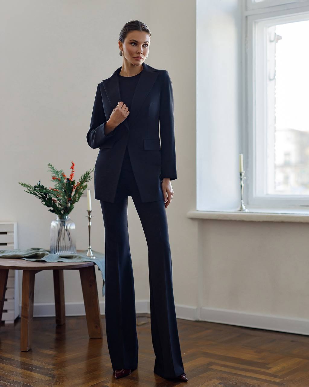 Woman wearing a navy blue suit standing in a room with a window and table.