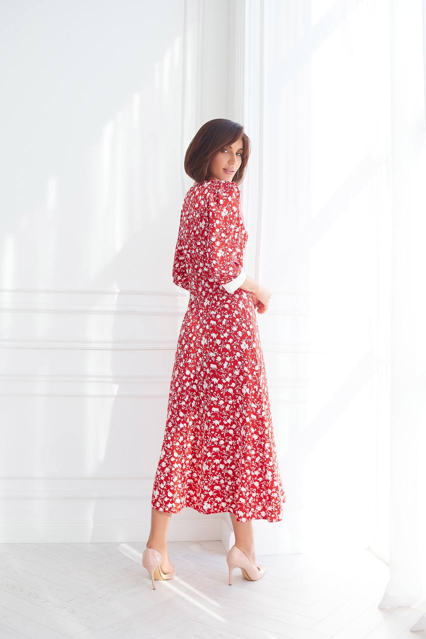 Woman wearing a red floral dress standing in front of white blinds.