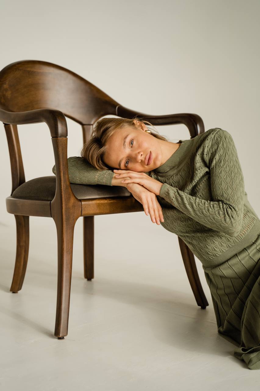 Woman in a green outfit lying on a wooden chair against a plain background