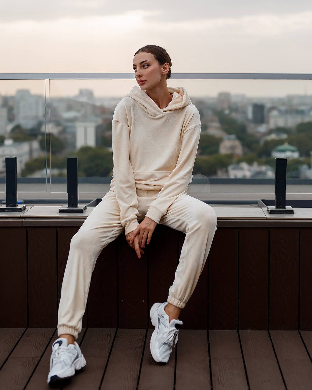 Woman in a beige tracksuit sitting on a rooftop with a cityscape in the background