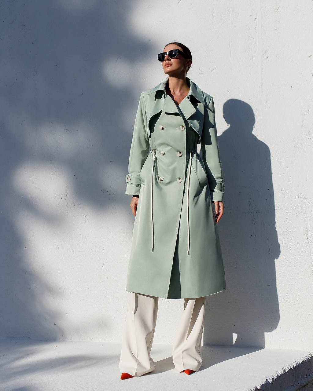Person wearing a long light green coat against a white wall
