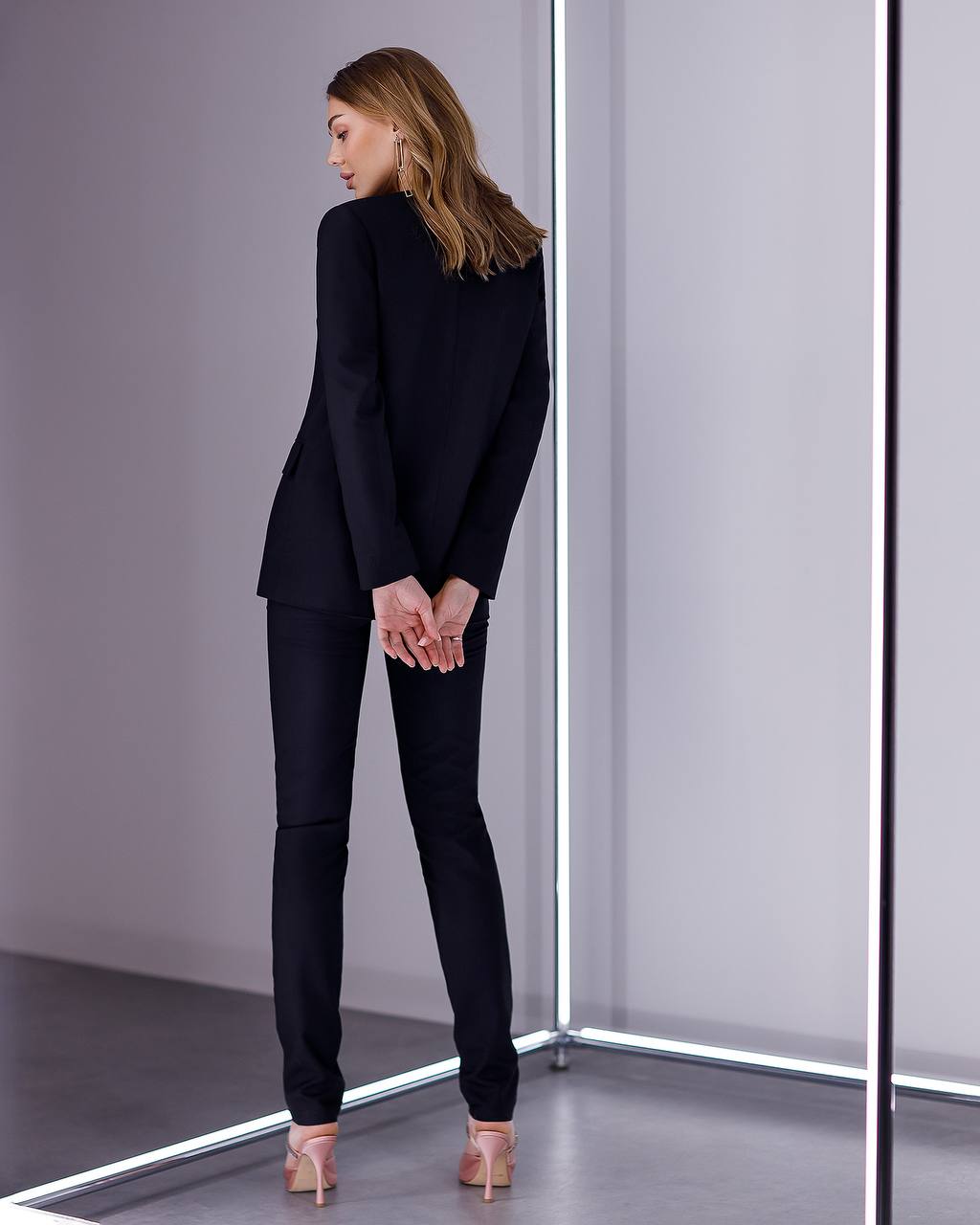 Woman in a black suit standing in front of a mirror in a minimalistic room.