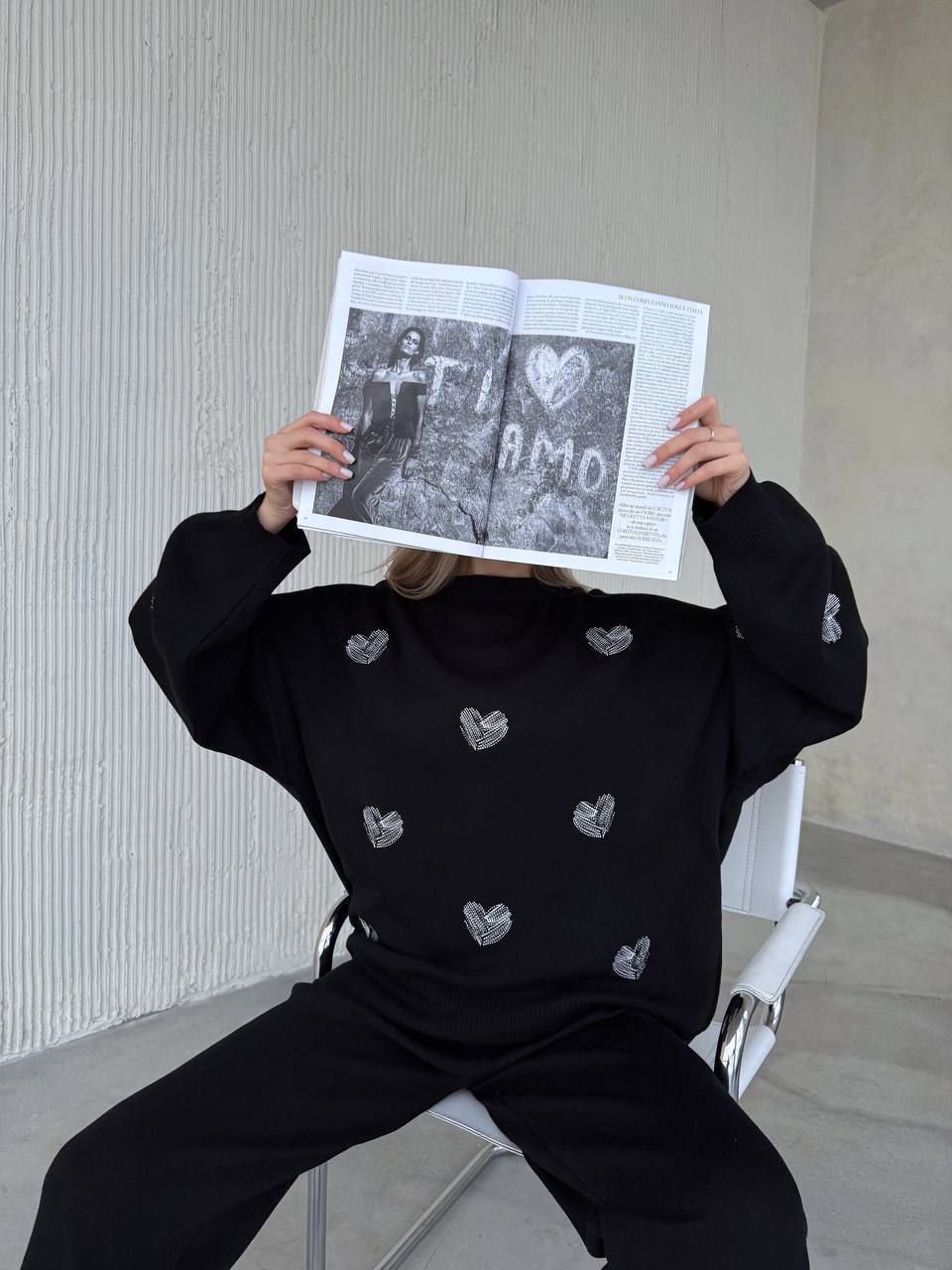 Person wearing a black sweater with heart patterns, holding a newspaper in front of their face.