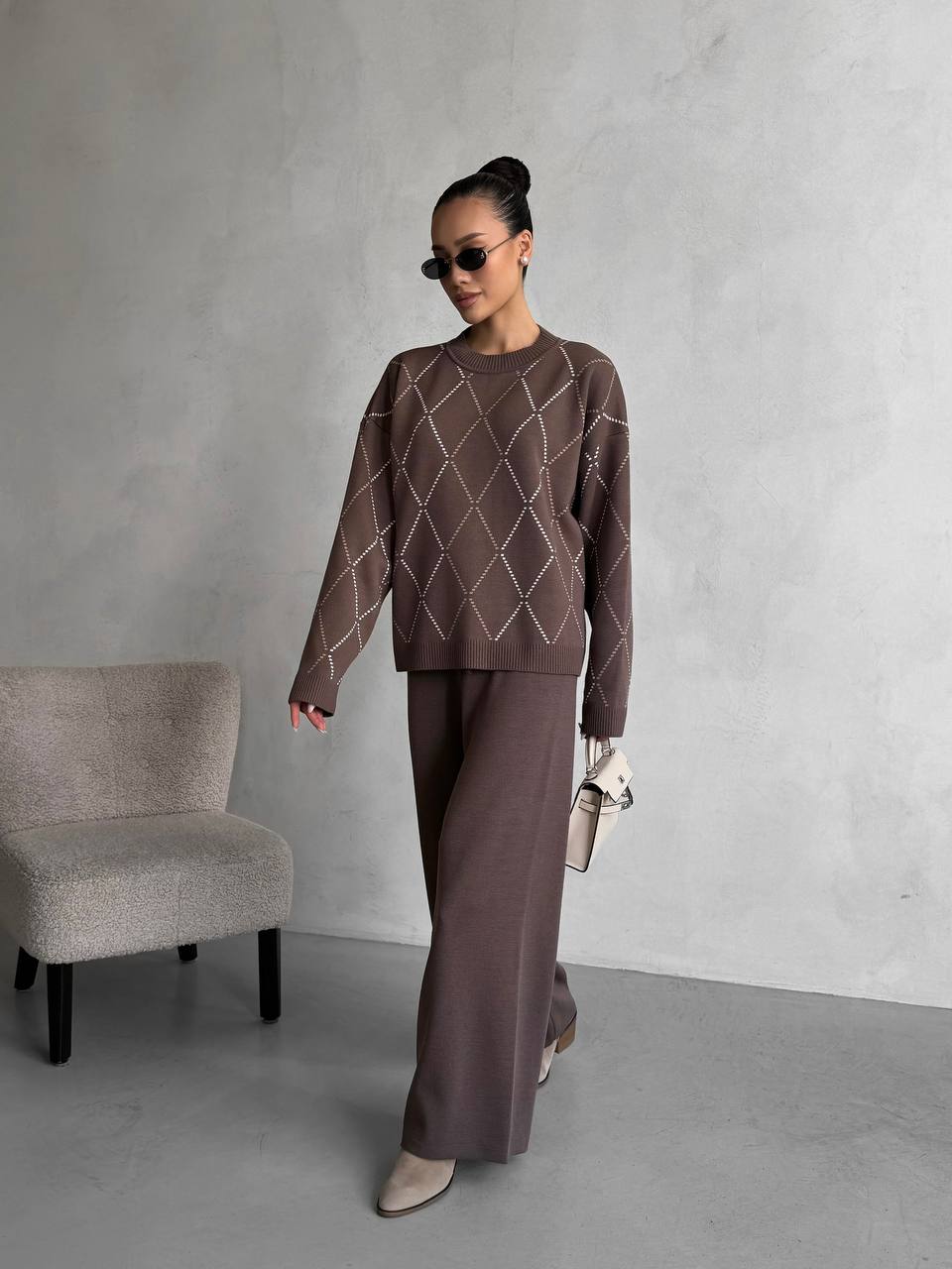Woman wearing a brown patterned sweater and pants standing against a gray wall.