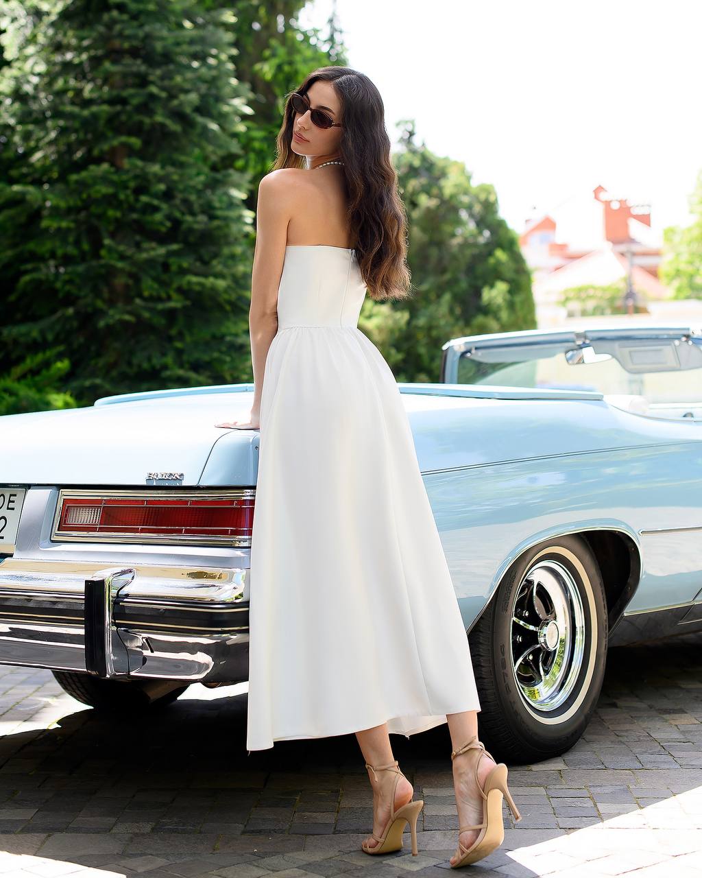 Woman in a white strapless dress standing next to a vintage car with trees in the background