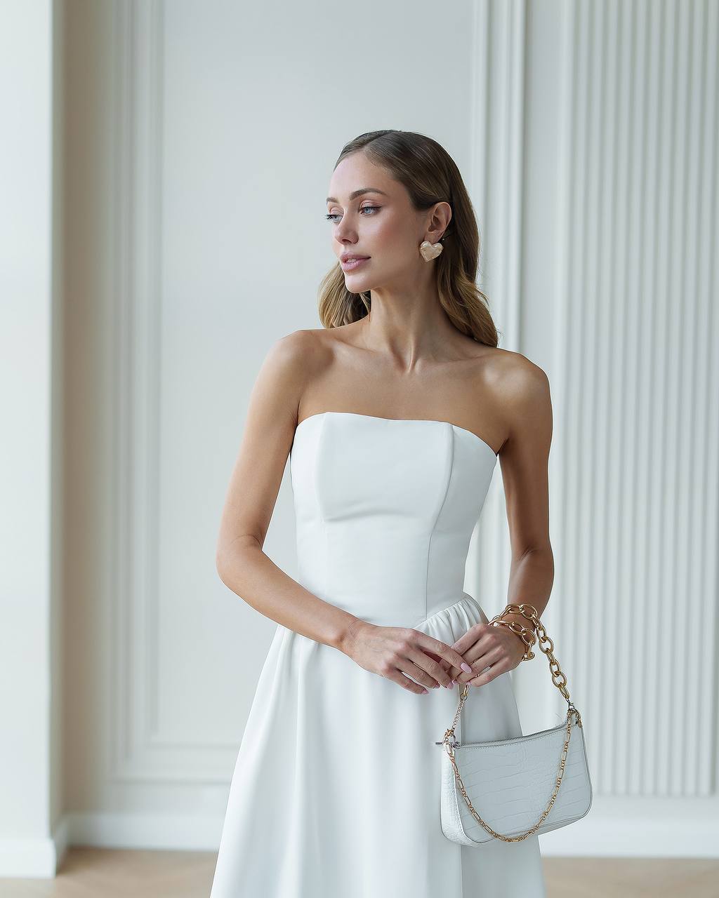Woman in a white strapless dress holding a white handbag with gold chain in a minimalistic setting.