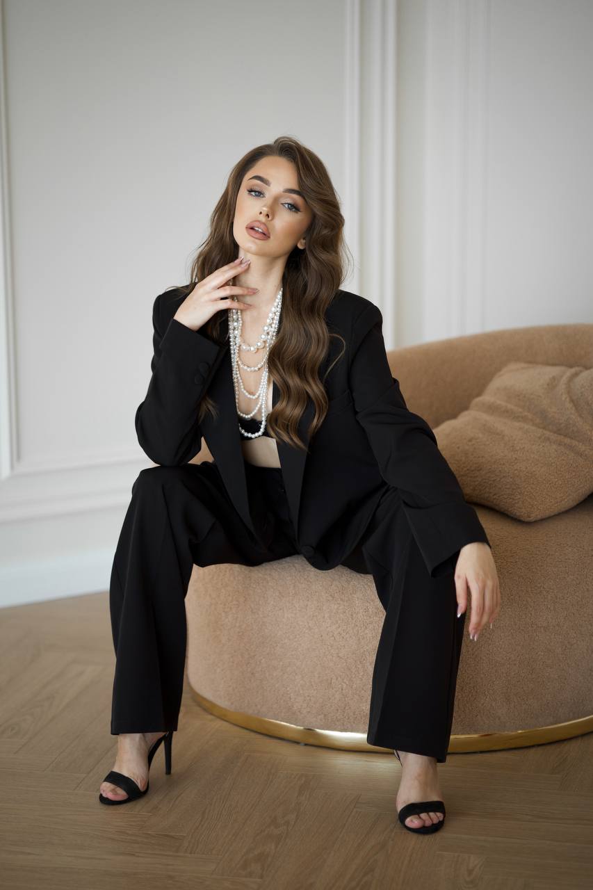 Woman in a black suit sitting on a brown couch in a room with white walls.