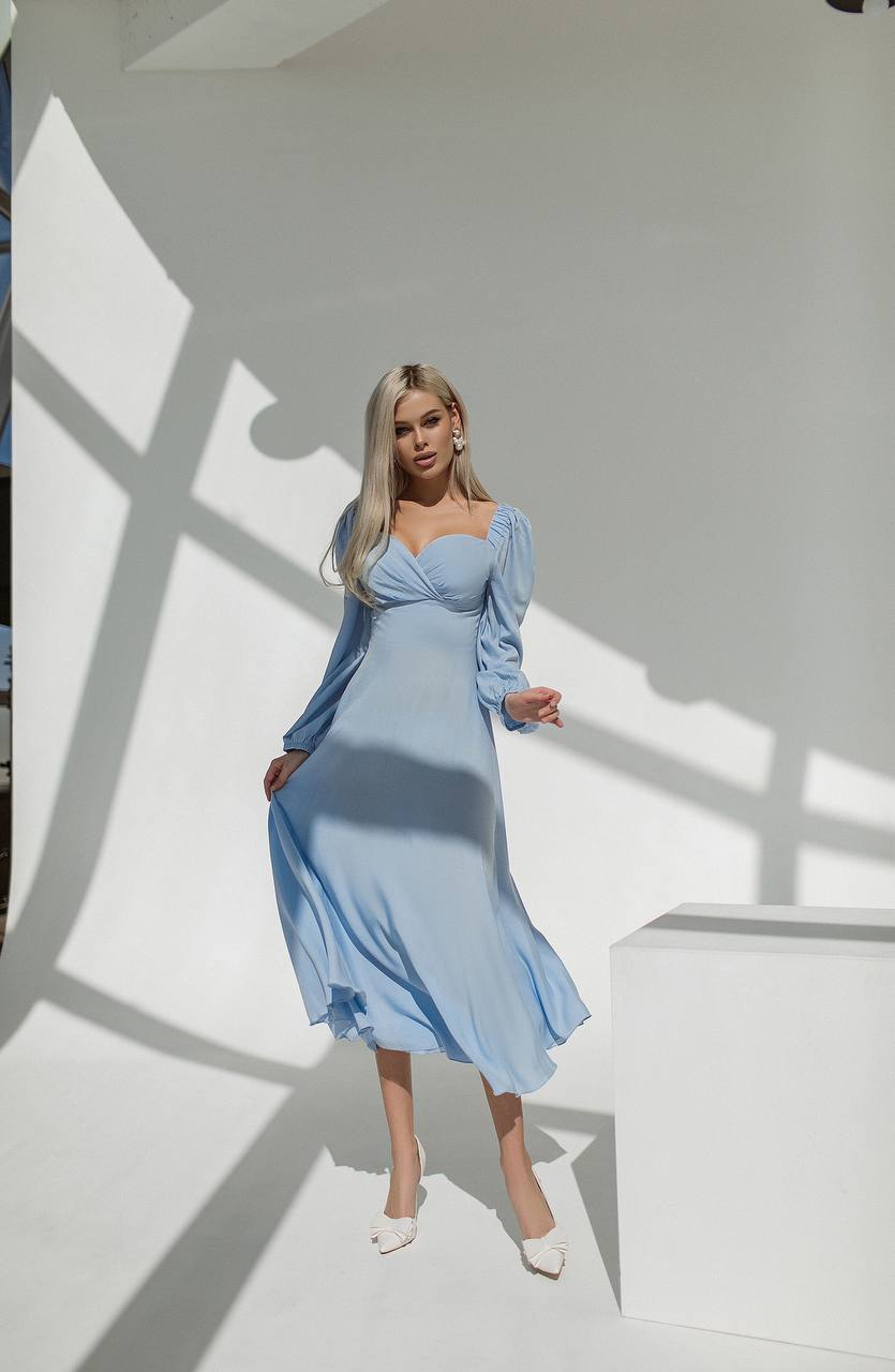 Woman in a light blue dress standing against a white wall with shadows.