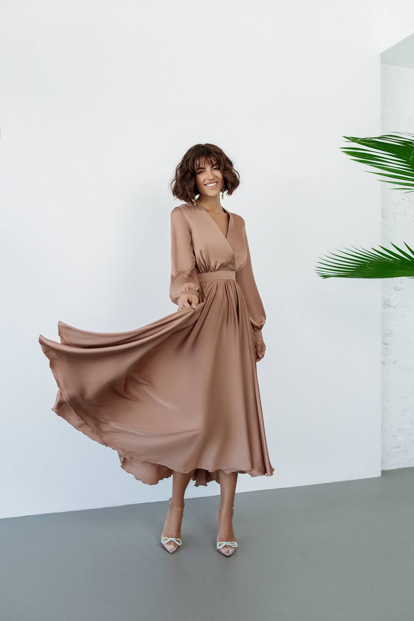 Woman in a flowing beige dress standing against a white background with a plant to the side.