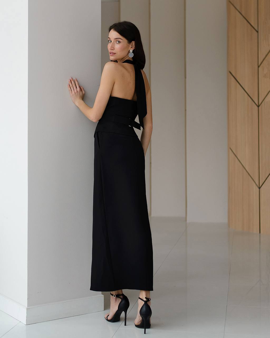 Woman in a black halter neck dress standing against a neutral wall.