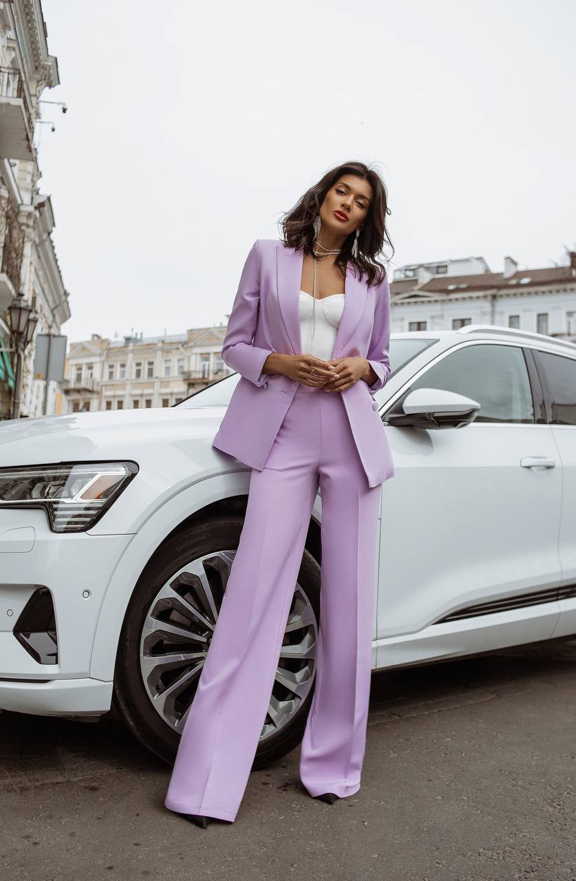 Woman in a lavender suit standing next to a white car on a city street.