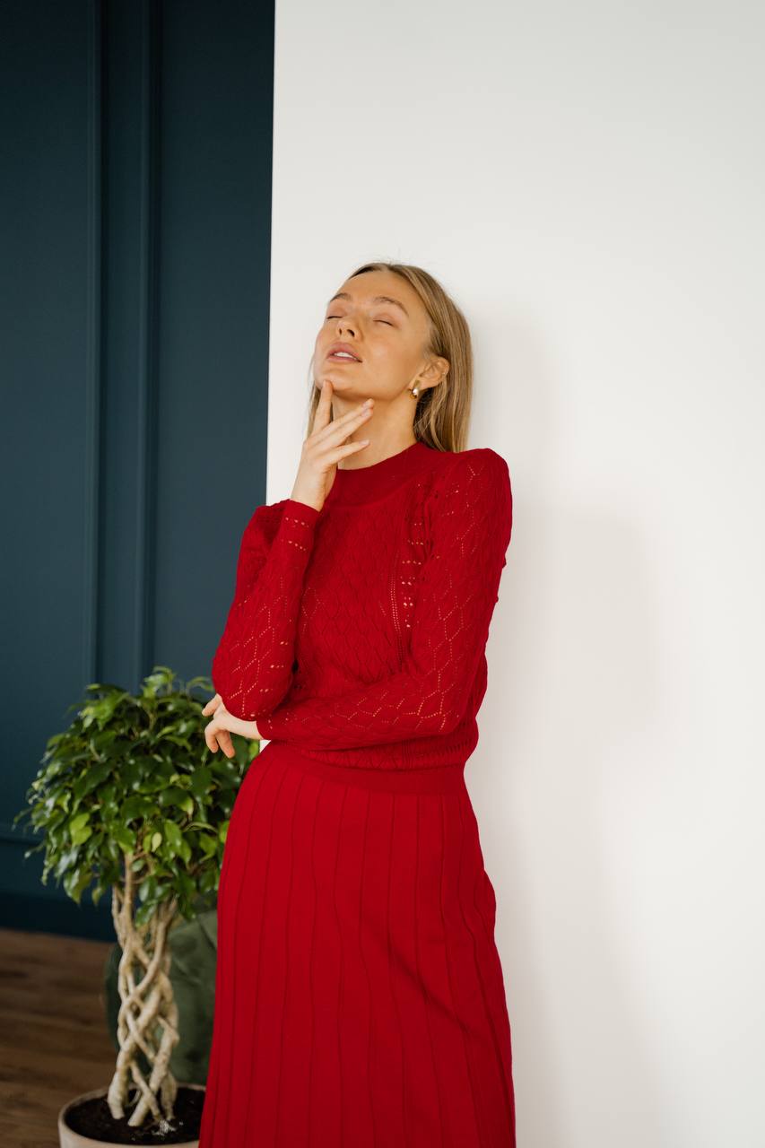 Woman wearing a red outfit standing against a white wall with a plant to her left.