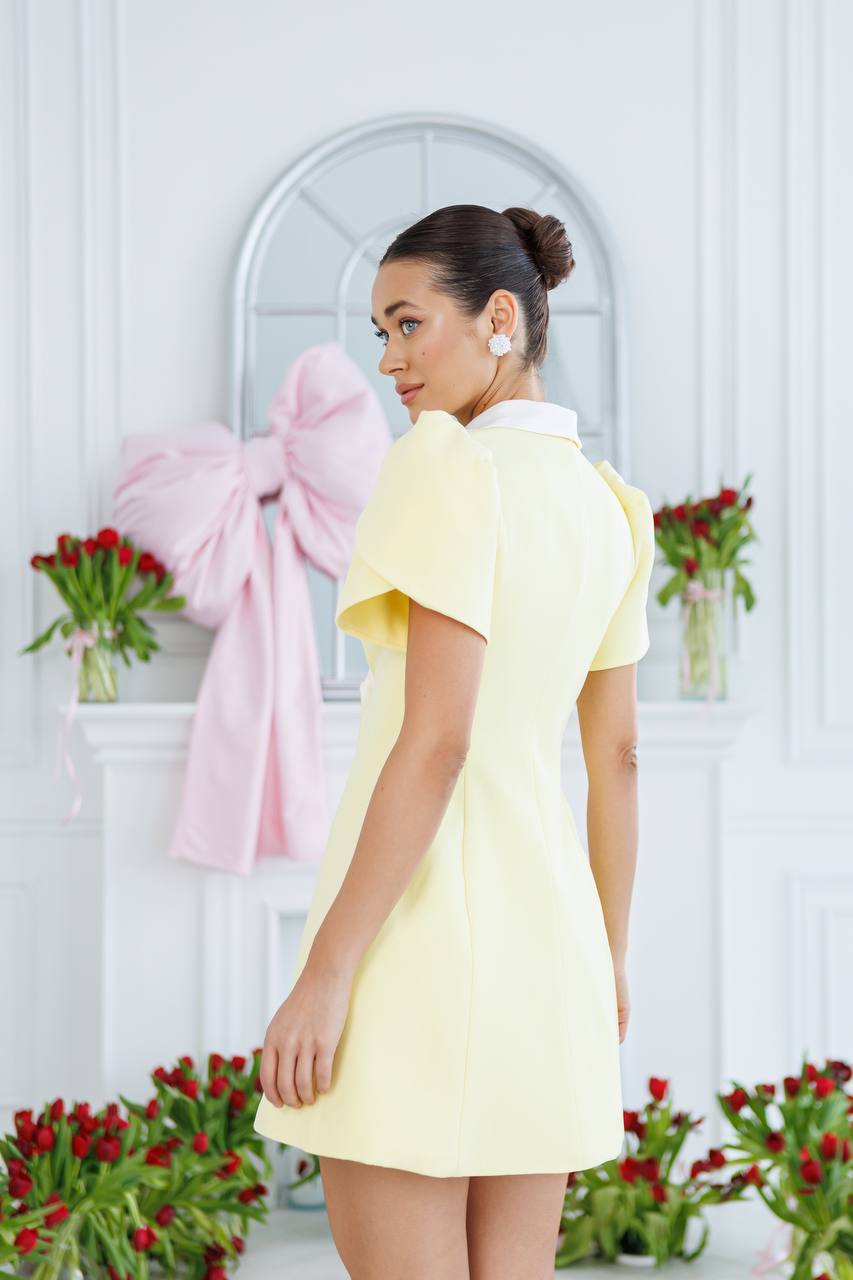 Woman in a yellow dress standing in front of a decorative wall with flowers and ribbons.