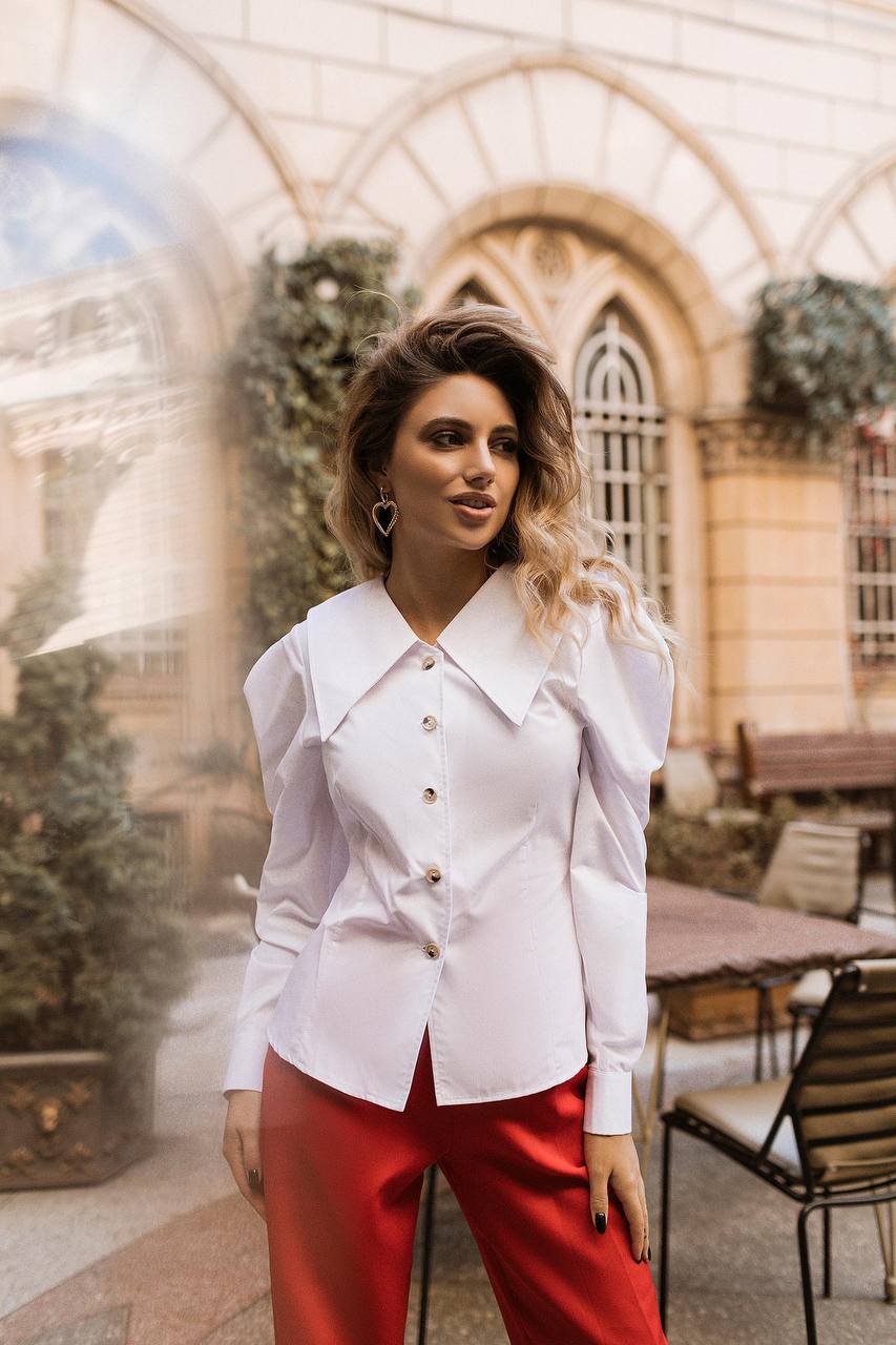 Woman wearing a white blouse and red pants standing in an outdoor setting with architectural elements.