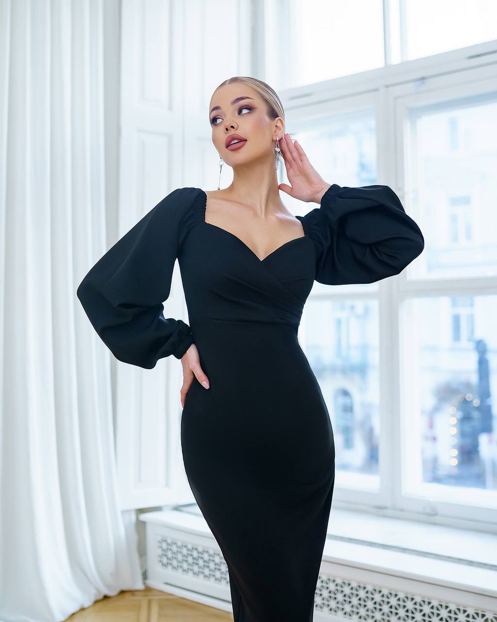 Woman in a black dress standing in a room with large windows.