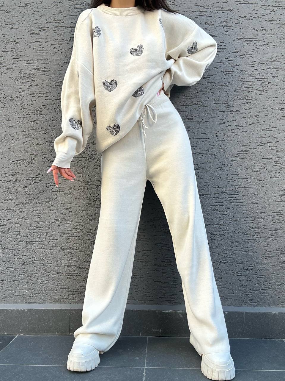 Person wearing a white outfit with heart patterns against a gray wall.