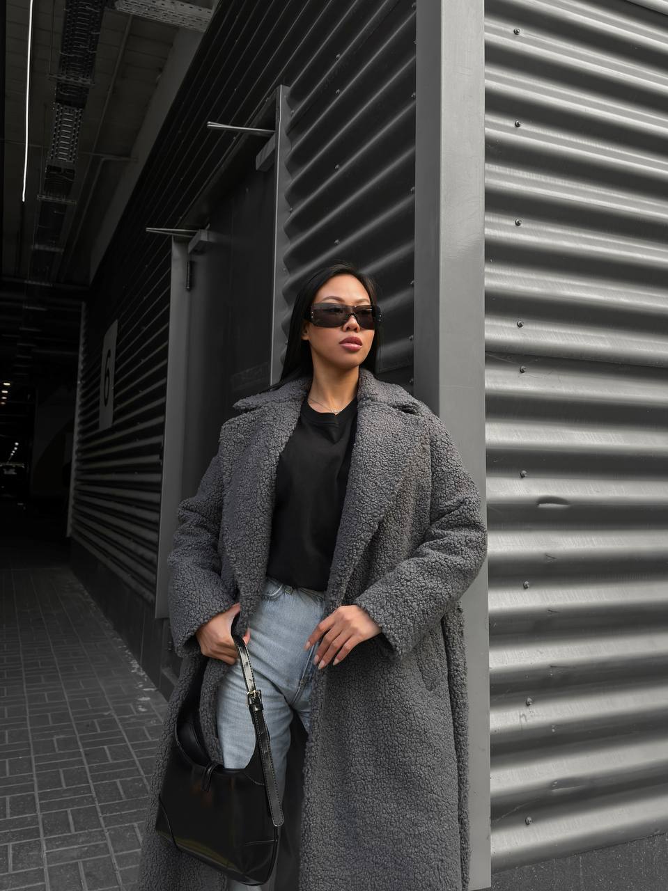 Person wearing a long gray coat and sunglasses standing against a corrugated metal wall.