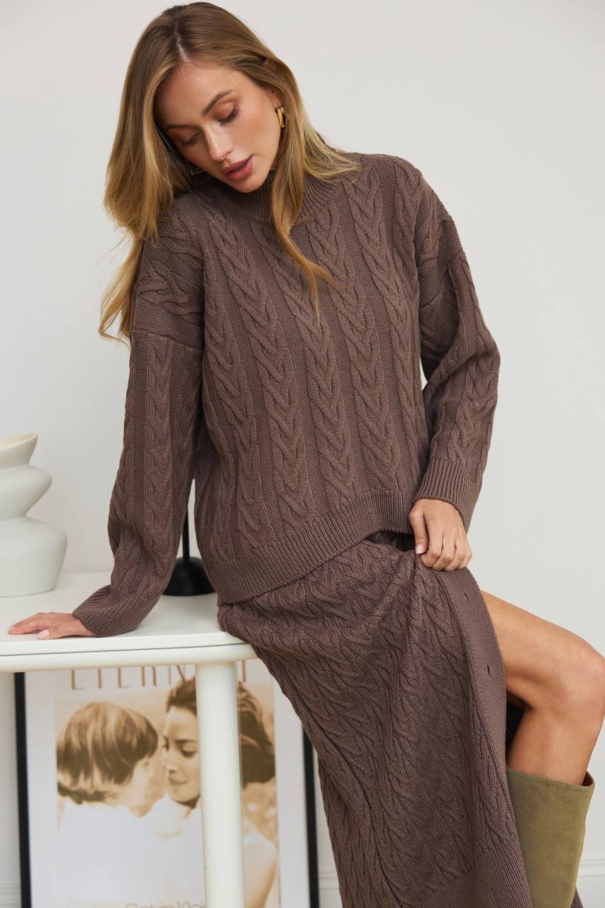 Woman wearing a brown cable knit sweater and pants sitting on a white surface.