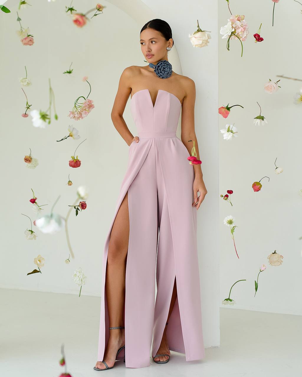 Woman wearing a pink strapless gown with a high slit, standing against a floral decorated wall.