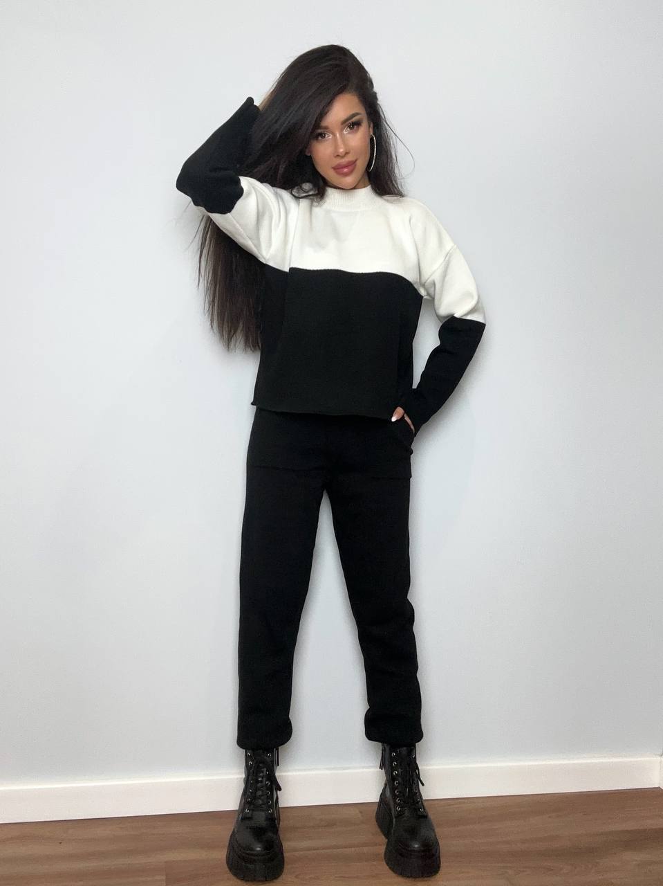 Person wearing a black and white outfit with black pants and boots against a plain wall.