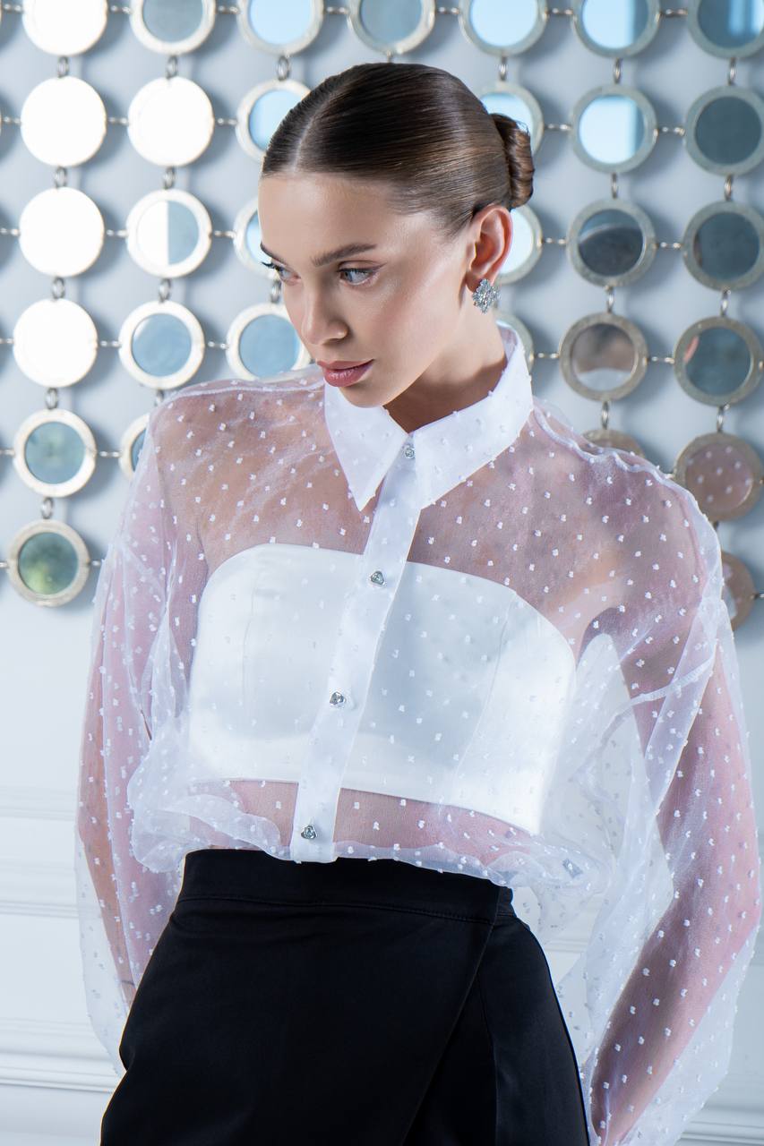Woman wearing a white blouse with sheer polka dot overlay against a decorative wall.