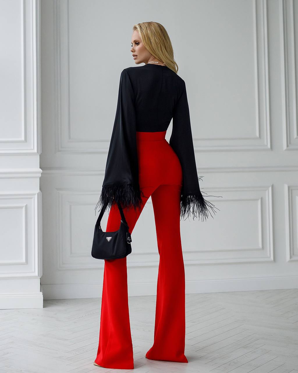 Woman wearing a black top with fringe and red pants, standing against a white paneled wall.
