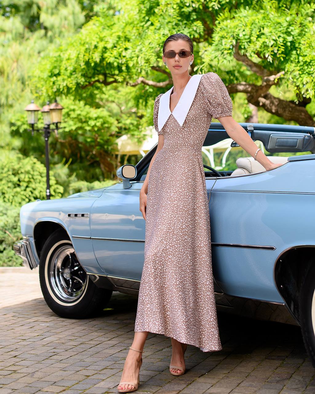 Woman in a patterned dress standing next to a vintage car with greenery in the background