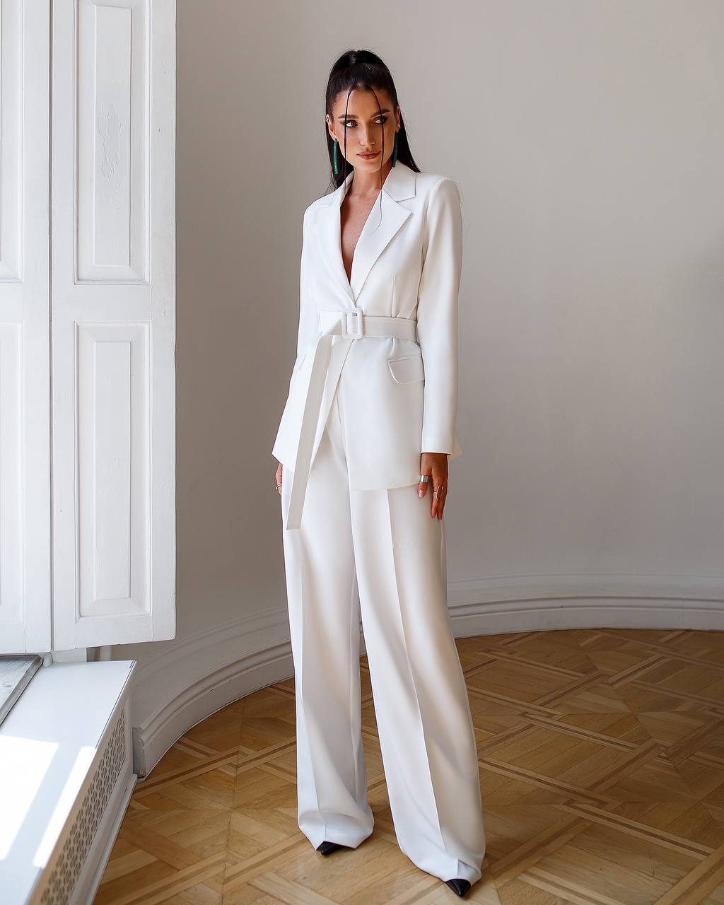 Woman wearing a white suit standing in a room with wooden flooring and white walls.