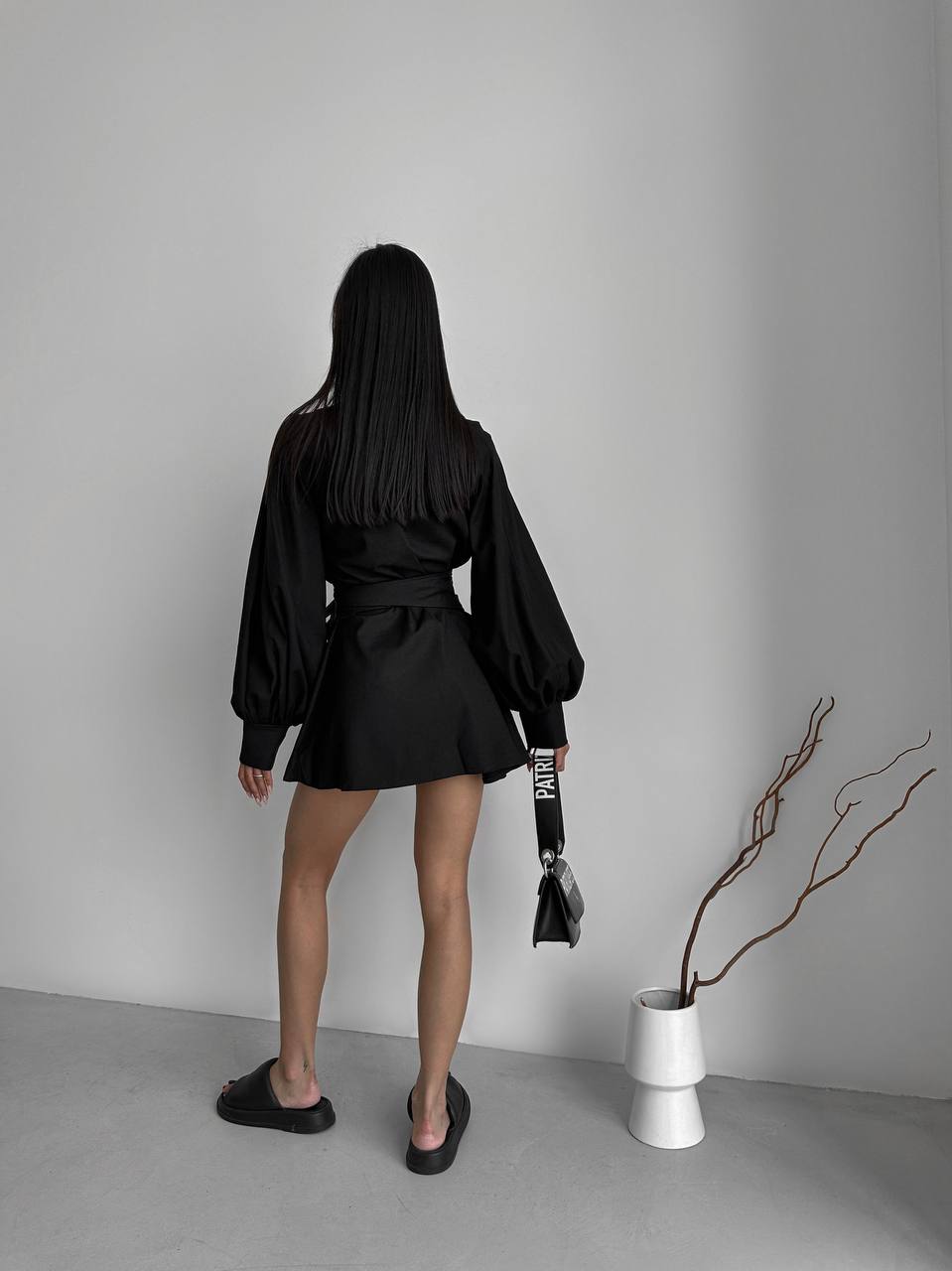 Person wearing a black outfit standing in a minimalistic room with a vase and branch.