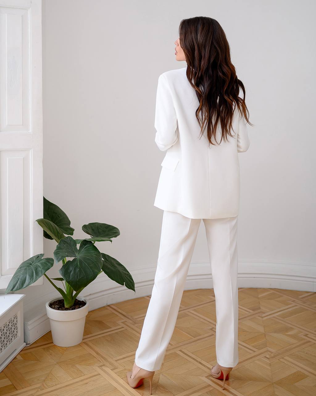 Woman in a white suit standing in a room with a plant and radiator.
