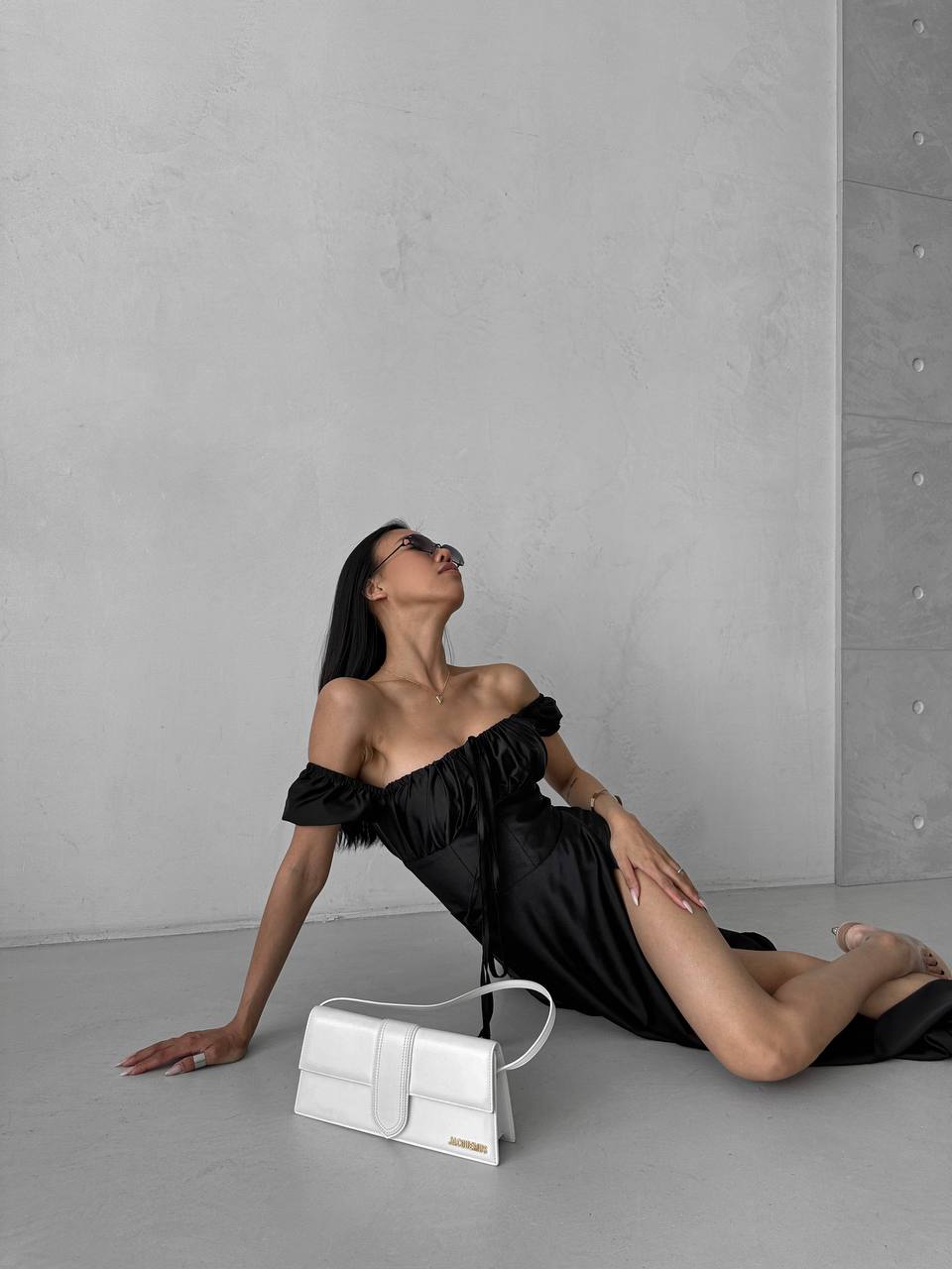 Woman in a black off-shoulder dress sitting on the floor with a white handbag next to her against a gray wall.