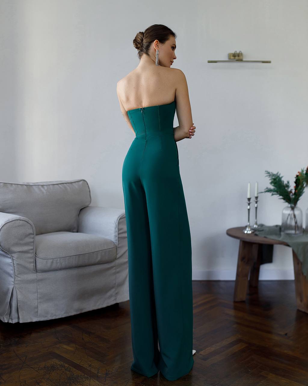 Woman in a strapless teal jumpsuit standing in a room with a gray sofa and wooden table.